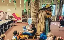 Bibliotekarka czyta dzieciom książkę pod bibliotecznym drzewem. 