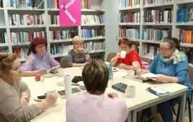 Przy stole siedzą uczestniczki spotkania i dyskutują. W tle regały z książkami i plakat DKK