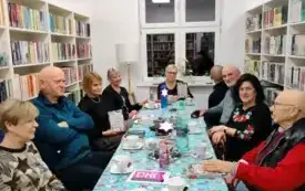 Uczestnicy Dyskusyjnego Klubu Książki siedzą przy stole, na kt&oacute;rym jest świąteczna cerata. Na stole leżą ozdoby, słodycze, filiżanki, talerzyki, książki, kartki. Dw&oacute;jka uczestnik&oacute;w spotkania trzyma książki w g&oacute;rze.