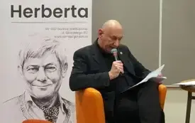 Robert Rudiak siedzi na pomarańczowym fotelu na tle baneru Biblioteki Herberta i odczytuje swoje wiersze do mikrofonu.