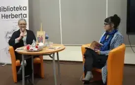 Agnieszka Kopaczyńska-Moskaluk oraz Beata Igielska siedzą przy udekorowanym  stole i omawiają zawartość czasopisma "Pegaz Lubuski". W tle widać baner Biblioteki Herberta.