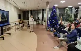 Po lewej stronie telewizor ze slajdem prezentacji Michała Gierkego, kt&oacute;ry m&oacute;wi do siedzącej w holu publiczności
