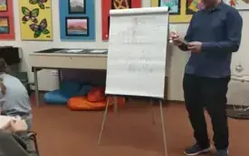 Prezenter rozwiązuje krzyż&oacute;wkę wraz z uczestnikami warsztat&oacute;w, w tle widoczny clipchart z hasłami.