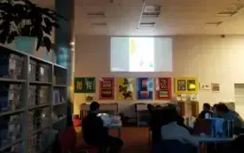 W pomieszczeniu panuje p&oacute;łmrok. Na ścianie widoczna prezentacja multimedialna. Uczestnicy warsztat&oacute;w siedzą na krzesłach.