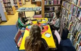 Przy stole kobieta i dwie dziewczynki czytają książki.