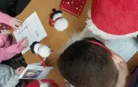 Jasno brązowy blat stołu, na nim kartki, skrzaty i czerwona koperta. Nad stołem pochylone głowy dzieci.