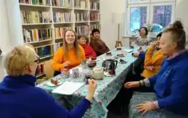 Uczestnicy spotkania siedzą przy stole i słuchają wykładu Małgorzaty Murowickiej-Kozickiej. Na stole są dzbanki z r&oacute;żnymi rodzajami herbat, talerzyki, słodycze. 