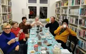 Uczestnicy spotkania edukacyjnego siedzą przy stole, na kt&oacute;rym są dzbanki z r&oacute;żnymi rodzajami herbat, talerzyki, słodycze. Uczestnicy piją herbatę z filiżanek. Po lewej i prawej stronie widać białe regały z książkami, a z tyłu białą ścianę i okno.