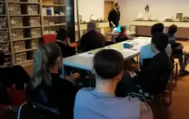 Na zdjęciu - w pomieszczeniu panuje p&oacute;łmrok. Uczestnicy warsztat&oacute;w siedzą przy stole. W tle prowadzący oraz wyświetlona na ścianie prezentacja multimedialna.