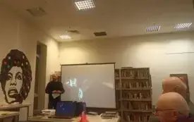 Uczestnicy siedzą przy stole, osoba prowadząca spotkanie stoi przy ekranie.
