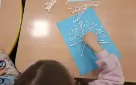 Dziewczynka układa patyczki higieniczne na niebieskiej kartce, tworząc płatek śniegu.