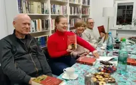 Na zdjęciu czw&oacute;rka uczestnik&oacute;w Dyskusyjnego Klubu Książki. Dw&oacute;jka z nich trzyma w g&oacute;rze książki Kristin Hannah &bdquo;Kobiety&rdquo;. Na stole są słodycze, napoje, filiżanki, talerzyki, logo DKK, książki. W tle widać okno i ciąg białych regał&oacute;w z książkami.