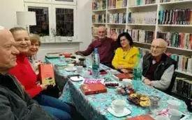 Uczestnicy Dyskusyjnego Klubu Książki siedzą przy stole. Na stole są słodycze, napoje, filiżanki, talerzyki, logo DKK, książki. W tle widać okno i ciąg białych regał&oacute;w z książkami. Moderatorka trzyma do g&oacute;ry książkę Kristin Hannah &bdquo;Kobiety&rdquo;.