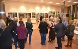 Na zdjęciu widać kilkanaście os&oacute;b stojących tyłem do aparatu, skupionych przed ścianą z obrazami i fotografiami. To wnętrze jasnej sali wystawowej z dużymi prostokątnymi lampami w suficie i błyszczącą, jasnobrązową podłogą. Ludzie stoją w kilku rzędach, w