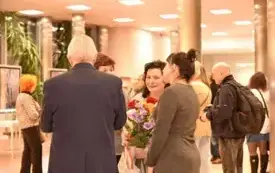 Grupa os&oacute;b w holu biblioteki. Na środku stoi kobieta z ciemnymi, kręconymi włosami do ramion, w jasnym stroju. Uśmiecha się i patrzy lekko w stronę aparatu. W obu rękach trzyma duży bukiet kolorowych kwiat&oacute;w &ndash; widać czerwone, fioletowe i r&oacute;żowe kwiaty z z
