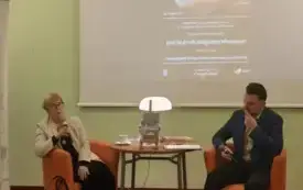 prof. Mikołajczak i dr Grzegorz Urbanek rozmawiają, siedząc na pomarańczowych fotelach przy okrągłym stoliku. Za nimi wyświetlony plakat wydarzenia