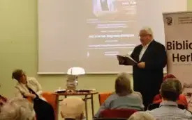 Dyrektor WiMBP Sławomir Szenwald przedstawia siedzącą na fotelu z lewej strony prof. Mikołajczak. Za nimi plakat wydarzenia, przed nimi uczestnicy zgromadzeni w sali
