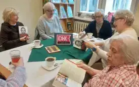 St&oacute;ł nakryty biało-zieloną serwetą, siedzące przy stole panie z otwartymi książkami, na stole, fioletowy emblemat DKK, filiżanki z napojami, czekoladki. W tle brązowa szafa z książkami, obok okno i parapet na nim doniczki z roślinami i r&oacute;żowo-czarna torba