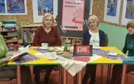 Czytelniczki dyskutują przy kawie i poczęstunku. Na stole położona jest jasna serweta i fioletowe serwetki.