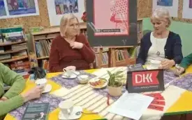 Cztery czytelniczki siedzą przy stole. Dyskutują o książce. Za nimi na sztaludze jest plakat Dyskusyjnego Klubu Książki. Na ściennie wiszą kolorowe prace plastyczne.