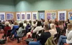Czytelnicy siedzą na krzesłach. Za nimi na ścianie są kolorowe prace plastyczne. Po lewej stronie jest plakat na sztaludze i pisarz z prowadzącą.