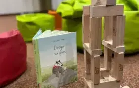 Wieża z drewnianych klock&oacute;w, obok stoi książka, w tle kolorowe pufy.