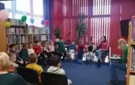 Kobieta czyta książkę grupie dzieci siedzących w p&oacute;łkole.