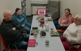 Na dole zdjęcia, w środku jest długi szary, w kształcie prostokąta st&oacute;ł. Po obu stronach stołu siedzą uczestnicy Dyskusyjnego Klubu Książki. Po prawej stronie dwie kobiety, po lewej stronie mężczyzna i kobieta. Na stole przed nimi leżą omawiane książki