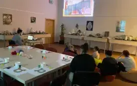 Prowadzący czyta informacje wyświetlane w postaci prezentacji multimedialnej na ekranie, w tle uczestnicy warsztat&oacute;w siedzący na kolorowych pufach.