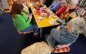 Dziewięć os&oacute;b siedzi przy stole, na pierwszym planie widoczny biały pies leżący przy stole.