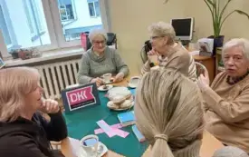 Siedzące przy stole panie, na zielonej serwecie książki, fioletowe emblematy DKK, filiżanki z napojami, pączki . W tle zielony kwiat , komputer, ż&oacute;łty kolor ścian i okno.
