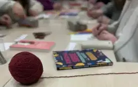 Kłębek bordowej wełny na długim stole, w tle widoczne dłonie dorosłych os&oacute;b. Książki leżą na stole.