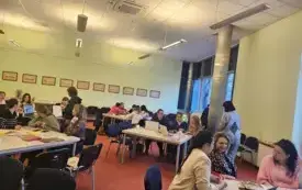 Uczestnicy szkolenia pracują w grupach przy komputerach. Trenerka porusza się między uczestnikami.