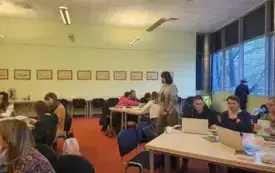 Uczestnicy szkolenia pracują w grupach przy komputerach. Trenerka porusza się między uczestnikami.