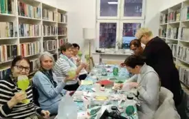 Uczestniczki zajęć siedzą przy stole. Jedna osoba stoi. Połowa uczestniczek siedząca po lewej stronie trzyma swoje świeczniki w g&oacute;rze prezentując je do zdjęcia. Pozostałe uczestniczki ozdabiają swoje świeczniki Na stole leżą serwetki, ozdoby, suszarka, fi
