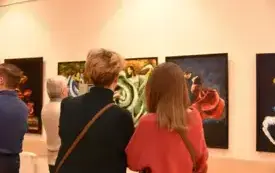 Dwie osoby stojące tyłem przed obrazami. Po lewej stronie stoi osoba z kr&oacute;tkimi, jasnobrązowymi włosami, ubrana w czarny golf; przez ramię przechodzi cienki, brązowy pasek torby. Po prawej stronie stoi osoba z prostymi, średniej długości włosami w kolorze