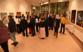 Grupa kilkudziesięciu os&oacute;b zgromadzona w przestronnym, jasnym holu.  Ludzie stoją w luźnych grupkach, rozmawiają ze sobą, część trzyma w dłoniach napoje.  Większość ma na sobie eleganckie lub p&oacute;łformalne ubrania w ciemniejszych kolorach &ndash; marynarki, koszu