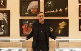 Artysta- Walenty Żemojtel stoi na tle swoich obraz&oacute;w. Ma  siwe włosy zaczesane do tyłu i wąsy, nosi okulary. Ubrany jest w  czarną marynarkę, czarną koszulę i ciemne spodnie, stoi wyprostowany z lekko rozchylonymi ramionami, oparte dłońmi o krawędzie dw&oacute;c