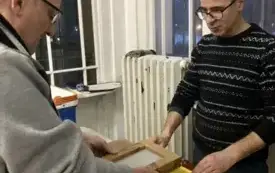 Dw&oacute;ch mężczyzn pracujących nad prostokątną drewnianą ramką nad ż&oacute;łtą plastikową miską z wodą - czerpią papier na sito.  Mężczyzna po prawej stronie ma jasną karnację, nosi czarne okulary i ma na sobie ciemny sweter w jasne, cienkie paski; stoi przodem do 