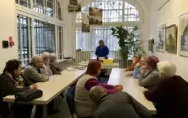 Zdjęcie grupy os&oacute;b siedzących przy długich stołach w jasnym pomieszczeniu.  W centrum, dalej od aparatu, stoi osoba w niebieskiej bluzce i zdaje się prowadzić spotkanie. Po obu stronach ustawione są stoły w kształt litery U, przy kt&oacute;rych siedzi kilkanaści
