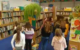Dzieci odnalazły w bibliotece dział z baśniami i stoją teraz między regałami. Bibliotekarka opowiada i pokazuje im r&oacute;żne książki.