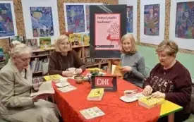 Cztery  czytelniczki siedzą przy stole. Rozmawiają o książce. Uśmiechają się. Za nimi wiszą kolorowe prace plastyczne.
