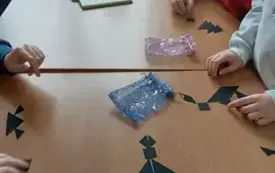 Na jasno-brązowym blacie otwarta książka, kolorowe woreczki, czarne figury geometryczne. Na blacie ręce dzieci.