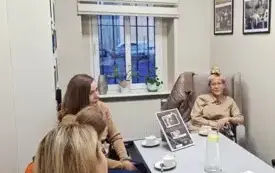 cztery kobiety siedzą przy stole, na stole znajdują się filiżanki do kawy, dzbanek na wodę i talerz ciasteczek. W tle widać okno i oprawione zdjęcia na ścianie