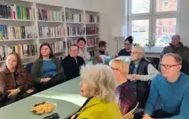 Uczestnicy spotkania &bdquo;Gorz&oacute;w znany i nieznany &ndash; tajemnicze zakątki Gorzowa Wielkopolskiego&rdquo; oglądają prezentację multimedialną o Gorzowie Wielkopolskim i słuchają prowadzącego, Zbigniewa Rudzińskiego. Na stole leży talerz ze słodyczami, okulary. Jedna 