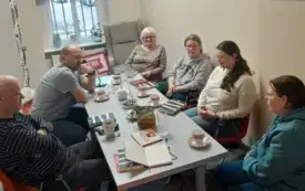 sześć dorosłych os&oacute;b, siedzących wok&oacute;ł długiego, szarego stołu w jasnym, kameralnym pomieszczeniu. Panuje atmosfera skupienia na rozmowie i wymiany refleksji na temat omawianej książki. Na stole przed klubowiczami rozłożone są liczne przedmioty, książki, 