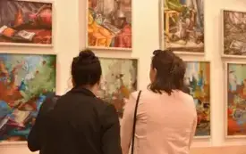 Dwie osoby stojące tyłem, oglądające obrazy na ścianie w galerii. Obie mają ciemne włosy; jedna ma włosy związane w kok i czarną g&oacute;rę, druga ma włosy spięte klamrą i jasnor&oacute;żową kurtkę. Na ścianie wisi kilka obraz&oacute;w w jasnych ramach, w intensywnych kolora
