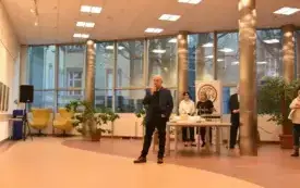 Grupa ludzi stojących tyłem w galerii sztuki przed ścianą pełną kolorowych obraz&oacute;w. Pomieszczenie jest jasne, z beżowymi ścianami i podłogą w ciepłym, brązowym odcieniu. Na ścianie wisi wiele niewielkich, barwnych prac ułożonych w rzędach. Dominują intens