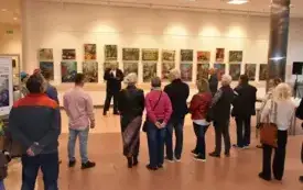 Duża sala wewnątrz budynku z wysokimi oknami na całej ścianie i kilkoma metalowymi kolumnami. W tle przez okna widać drzewa i zabudowania na zewnątrz. Na środku stoi długi st&oacute;ł z kilkoma osobami za nim. Po lewej stronie na pierwszym planie stoi mężczyzna 