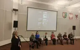 Na scenie w auli znajdują się: prowadząca Agnieszka Kopaczyńska-Moskaluk oraz goście spotkania siedzący na krzesłach. Za nimi na projektorze wyświetlony jest plakat wydarzenia
